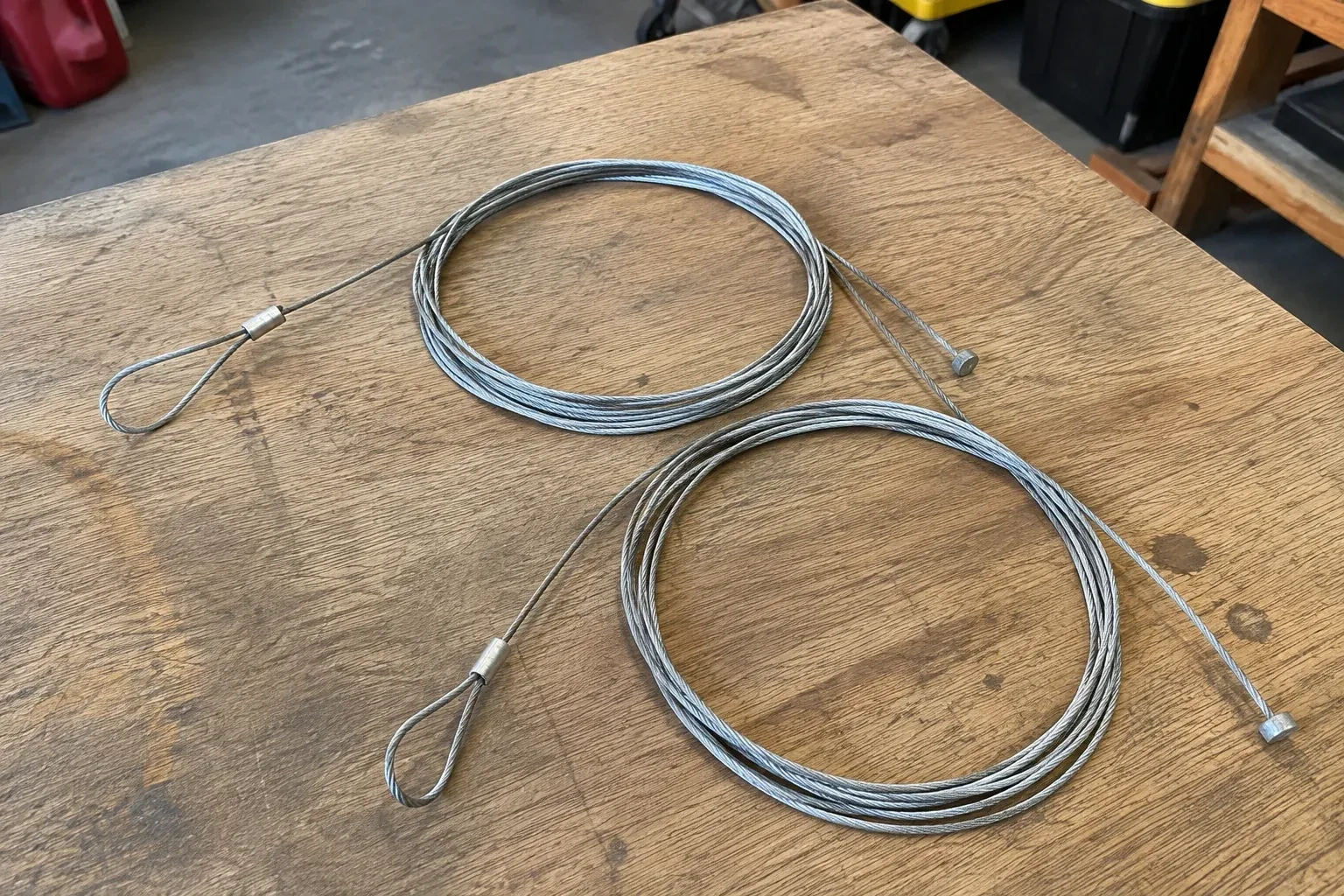 Two coiled steel cables with loop and stop ends on a workbench. — Garage Door Cable Replacement, Cundles