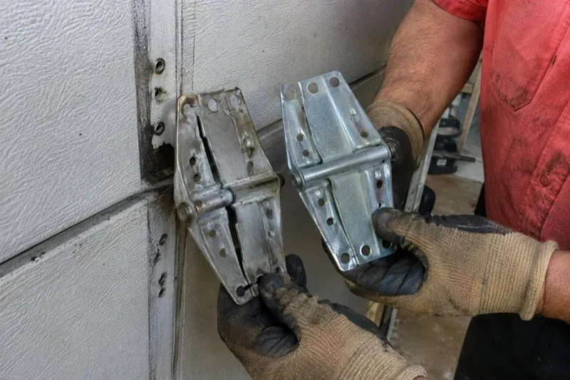 Gloved hands holding two metal garage-door hinges side by side, one discolored with a visible crack and the other bright silver. — Garage Door Hinge Replacement, Barrie