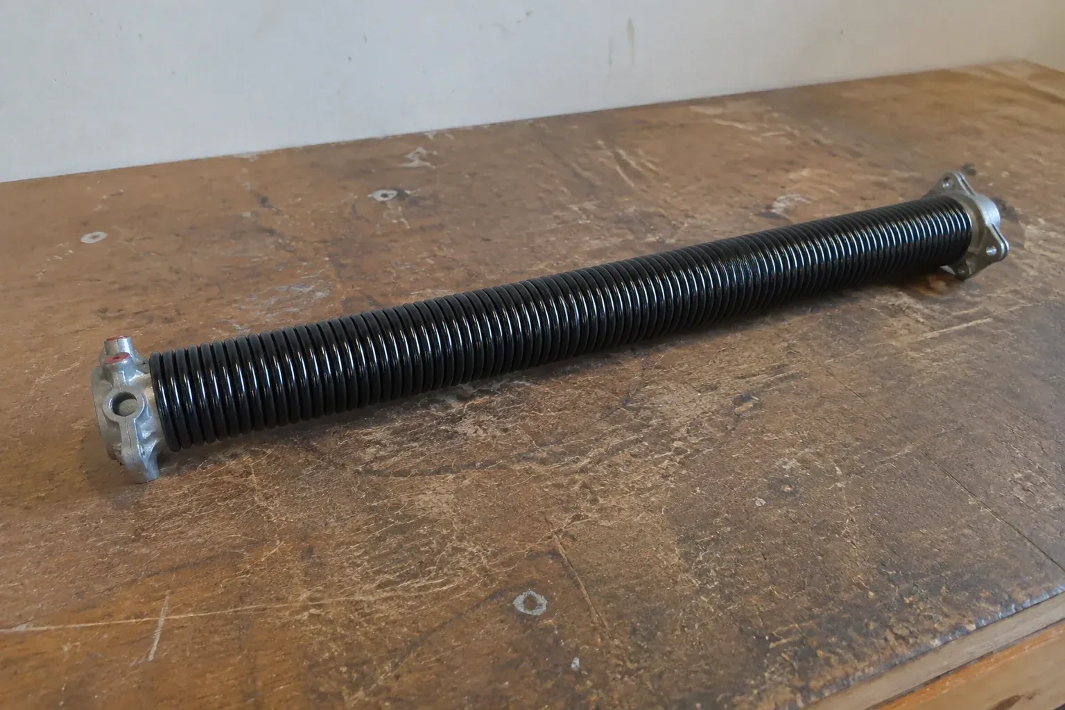 Detached black coil spring with metal end fittings on a workbench. — Torsion Spring Replacement, Cundles